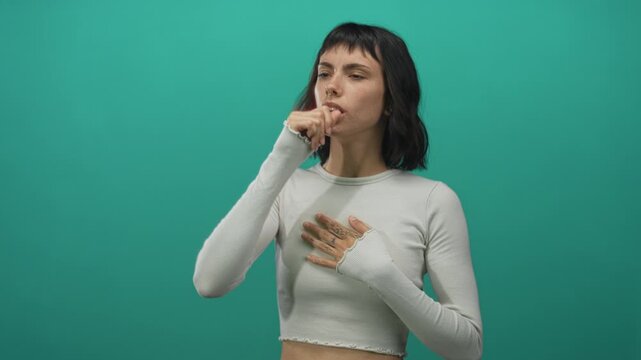 Young hispanic woman coughs with hand on chest and eyes closed against green backdrop in studio; concern.