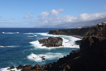 Wild Atlantic waves crashing against jagged volcanic outcrops on Tenerife&rsquo;s northern coast with distant mountains and dramatic sky