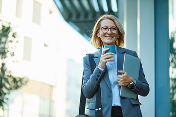 Fototapeta premium Portrait of a middle aged mature businesswoman woman using a laptop computer and holding a cup of coffee in a city park, surrounded by modern corporate office buildings