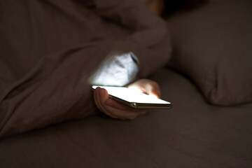 Female hand holding smartphone glowing under blanket in dark bedroom setting. © elenavolf