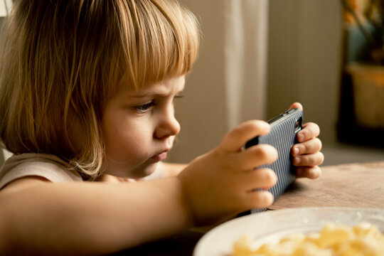 Little Child Girl using Phone while eating Pasta during Lunch at Table. 3 Years Toddler Kid Eating Food and Watch Cartoon on Smartphone. Nomophobia or Attention Deficit Hyperactivity Disorder ADHD