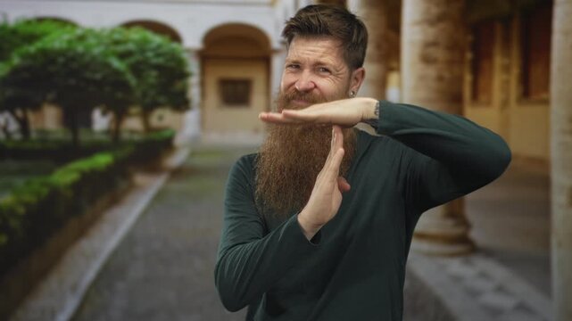 Man with long red beard makes time out gesture with hands on cobblestone street beside historic arches; pause.