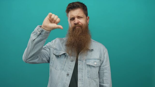 Young caucasian bearded man shows thumbs down gesture in teal studio; rejection frustration dissatisfaction critique.