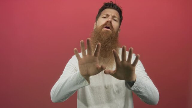 Man with long beard wearing white shirt pushes both palms forward toward camera in studio setting; resistance.