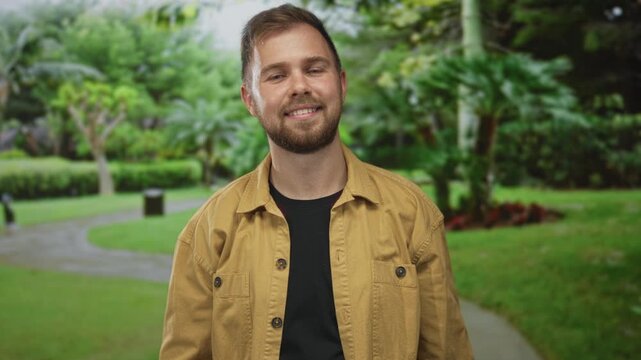 Man with beard smiles with eyes closed and points finger to camera with right hand while wearing a tan jacket on a forest path lined with green trees; confidence invitation.