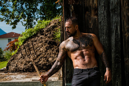 A strong sexy farmer poses by the farm. Muscular man holds a pitchfork. Rural life. Rural man works by the barn. Countryside lifestyle. Hard farm labor. Rural lifestyle. Farm worker holds a pitchfork.