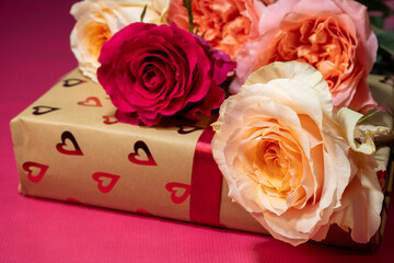 Fototapeta premium a gold gift box wrapped in heart-patterned paper and a red ribbon, topped with a blooming red rose and several cream and peach roses on a pink background.