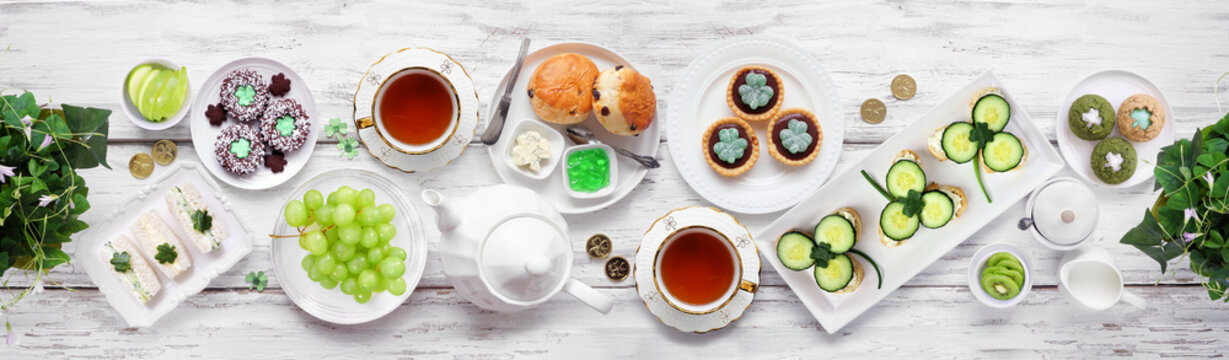 St Patrick's Day tea party table scene against a white wood banner background. Variety of green shamrock theme finger sandwiches, fruits, scones and sweet desserts. Overhead view.