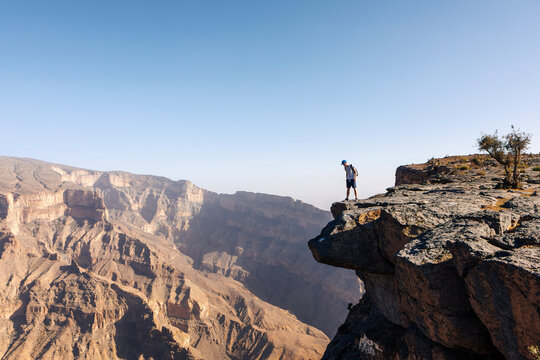 Tourist with backpack standing on cliff edge overlooking deep canyon in Jebel Akhdar, Oman. Dramatic landscape and sense of scale representing adventure travel, exploration, and wilderness.