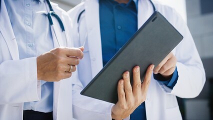 Close view of two doctors standing outside clinic. Medical specialists using tablet while reviewing symptoms. Focus on coordinated teamwork. Hands pointing at screen during case evaluation.