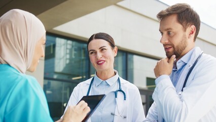 Female nurse holding tablet while asking questions. Smiling doctor focusing on details while listening. Male doctor explaining treatment steps carefully. Standing outside large hospital building.