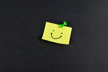 Yellow sticky note with smiley face pinned by green pushpin on black background, conveying positivity through simple, bold contrast.