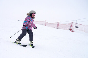 A little girl wearing a protective helmet is skiing down a snowy slope. She has ski poles in her hands.