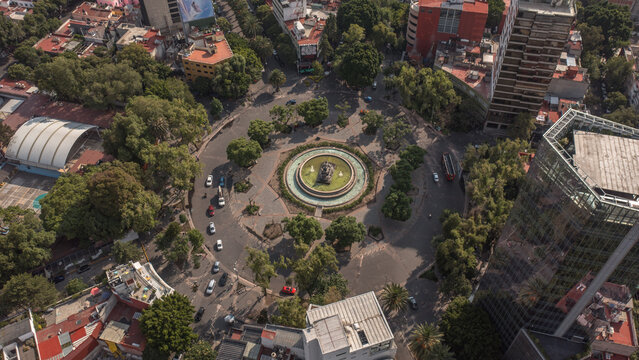 Aerial view of the Fuente de Cibeles roundabout in the Roma neighborhood of Mexico City. Capturing the iconic fountain surrounded by tree-lined streets, urban traffic, and residential buildings.