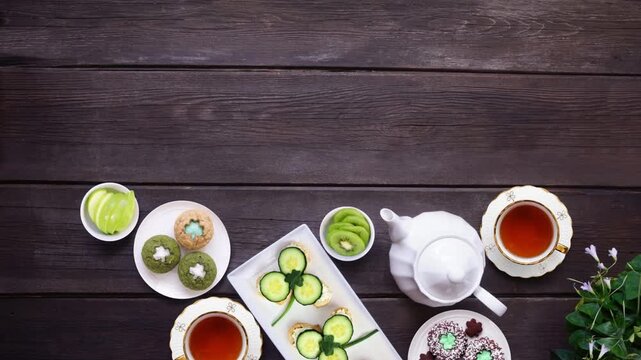 St Patrick's Day tea party bottom border table setting over a dark wood background. Stop motion video. Variety of green shamrock theme finger sandwiches, fruits, scones and sweet desserts. Top view.