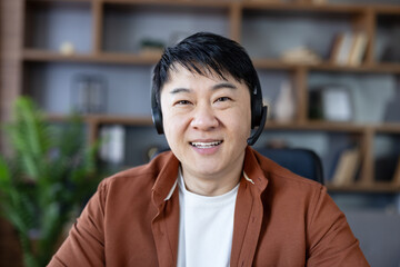 Fototapeta premium Asian man smiling, wearing a headset with a microphone, participating in an online video conference or remote consultation from a home office environment