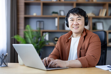 Fototapeta premium Smiling asian man wearing headphones, sitting at a desk, typing on a laptop, and working from a home office, embodying remote work and comfortable productivity