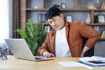 Fototapeta premium Asian man experiencing severe lower back pain while sitting at a desk and working on his laptop in a home office setting, indicating health issues from prolonged sitting