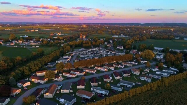 A sunset lights up a small town of mobile, manufactured homes surrounded by fields. The sky shows bright colors as evening falls. People relax in yards and enjoy the scenery.