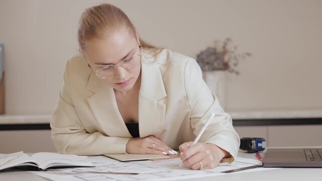 Caucasian female architect reviewing blueprints at office desk with computer.