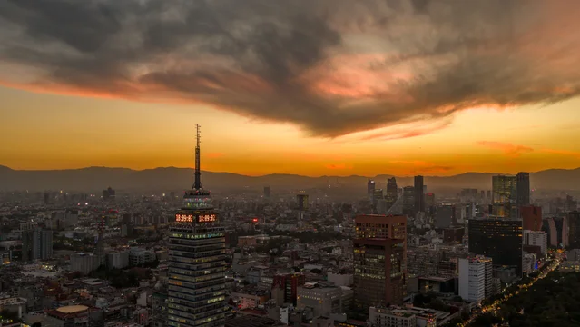 Aerial view of Mexico City at sunset featuring the Torre Latinoamericana and Paseo de la Reforma. Warm evening light over the urban skyline with dramatic clouds and city buildings.