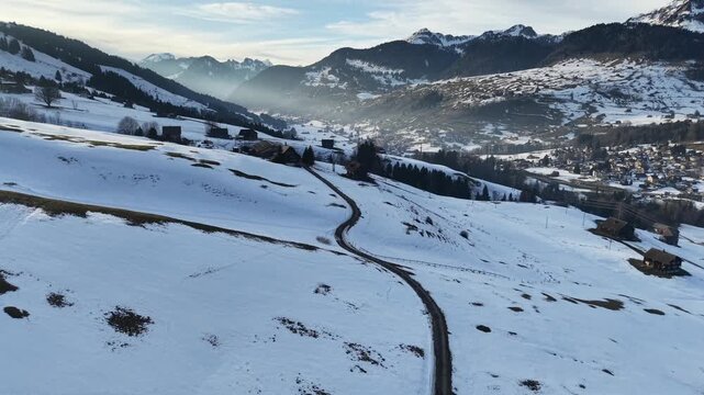 Drone flight forward over a cleared mountain road with patchy snow and occasional grass, surrounded by alpine slopes, distant mountain peaks visible, winter scenery in the Swiss Alps.