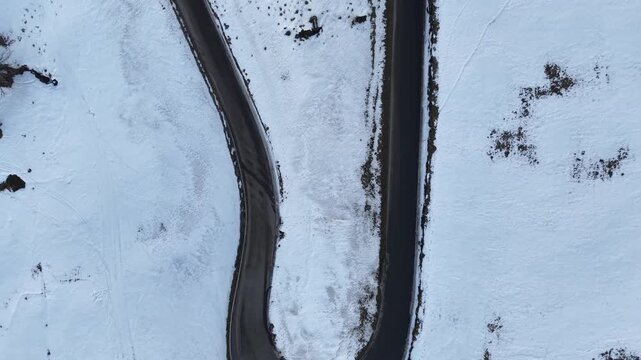 Top-down drone flight over a hairpin 180&deg; mountain road, ascending while moving forward, cleared asphalt with patchy snow along the edges, Swiss Alps winter landscape.