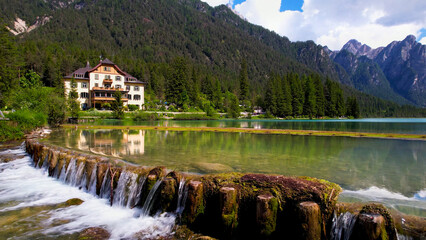 Obraz premium Lake Dobbiaco with Waterfall: Traditional House and Mountains View at Water Level, Italian Dolomites