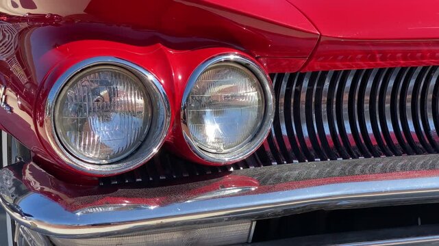 Bright red classic car front shows two round headlights, chrome trim and a sculpted hood. Glossy paint and curved grille lines emphasize the vintage automobile design.