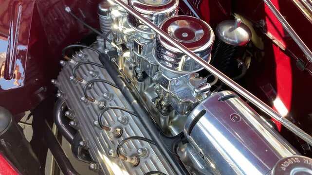 Closeup of a classic car engine shows three shiny chrome carburetors under the open hood. Sunlight reflects off polished metal parts, highlighting the detailed vintage performance motor.