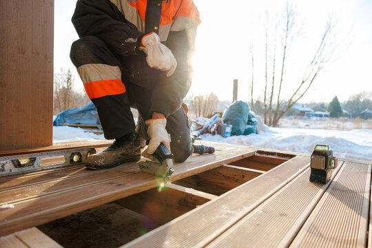 A worker in an orange and black jacket kneels on a wooden deck, uses a nailer, spirit level, cordless drill, and green laser level as sunlight flares through bare trees and snow.