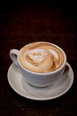 Fototapeta premium White ceramic cup of coffee with milk foam swirl on saucer, placed on dark wooden table, close view of hot cafe drink.