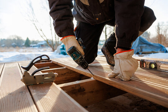 A tradesperson in gloves kneels on timber joists, driving screws into composite decking as a level and staple gun rest nearby on a cold day with snow and bare trees.