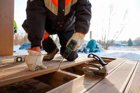 A deck installer kneels on joists, driving a screw with a cordless drill into composite boards. Snow lies beyond, tools rest nearby. Warm afternoon light marks cold season work.