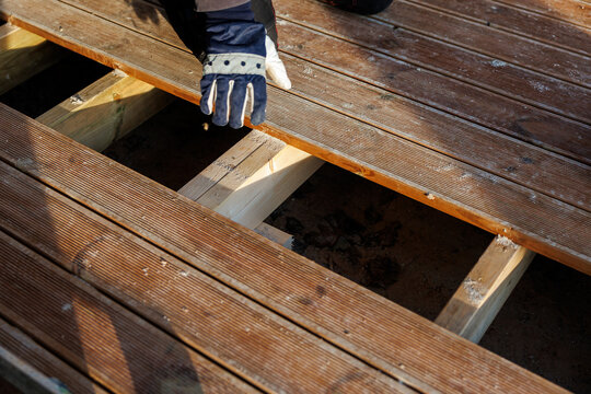 A gloved worker positions a grooved timber plank on joists, with a gap revealing substructure and fresh supports, sawdust present, close up view of alignment process.