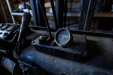 Close view of a weathered round gauge with a chipped blue rim on a grime coated forklift dashboard, beside rusted levers and a taped gear stick in an indoor workshop. © True Pixel Art