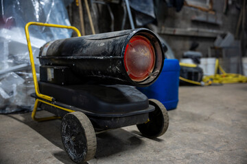 Black wheeled torpedo heater with yellow handle sits on concrete floor, burner glowing red, tools and plastic sheeting nearby, blue fuel container and coiled cords present.