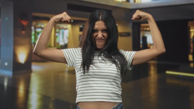 Woman flexes arms and grimaces while showing bare midriff in a building lobby; confidence empowerment.