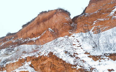 Eroded ravine wall revealing striking clay layers, topped with snow and dangling grass roots.