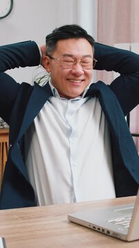 Asian businessman freelancer taking break leaning on chair after working celebrating success victory at modern home office workplace desk. Male Chinese in formal suit puts hands behind head relaxing