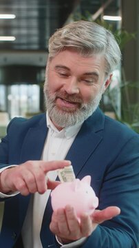 Happy mature businessman entrepreneur save money using pink piggybank putting dollar cast inside. Success, economy, investment, deposit. Bearded guy in suit sitting with laptop in modern office lobby.