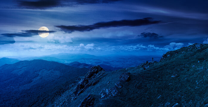 breathtaking summer mountain landscape at night. rocks on edge of hill in full moon light. view in to valley. grassy slope under cloudy sky. distant ridge on the horizon. epic mysterious fantasy