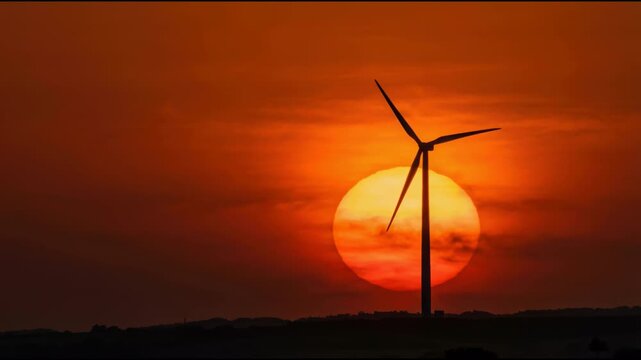 Cinematic silhouette of wind turbine rotating slowly against large orange setting sun. Dramatic golden hour sky. Solar and wind power generation concept. Global warming solution.