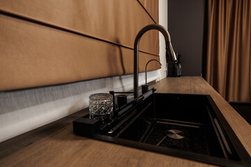 Close up of matte black inset sink and arched bronze faucet, with crystal cut tumbler and soap in a...