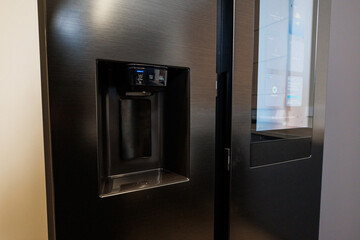 Matte black stainless steel fridge shows water and ice dispenser and a glass touchscreen with widgets and controls. Soft indoor lighting highlights brushed metal and clean lines.