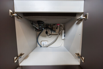 Open kitchen sink cabinet shows drain trap, flexible hoses, right side water filter, two socket...
