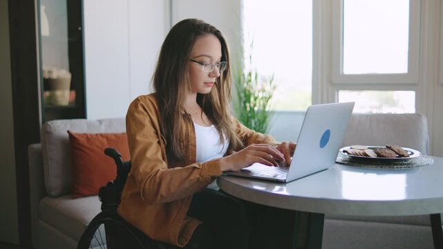 Woman remote working at home using laptop at table in bright room during daytime while sitting in wheelchair