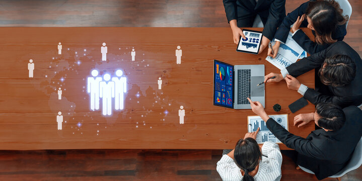 A diverse team collaborates around a wooden table, utilizing digital analytics and business graphics to enhance strategy and productivity in a modern office environment. Trope - Powered by Adobe