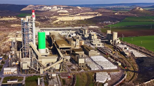Massive industrial site includes large buildings, stacks, and equipment located near green fields and hills under clear sky. Train tracks run alongside the facility with vehicles present.