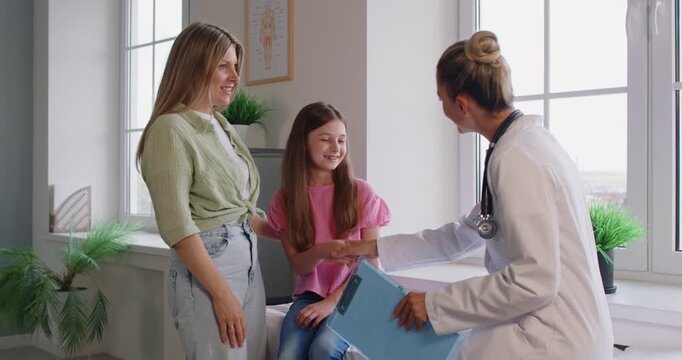 Mother and daughter meeting pediatrician for consultation, routine checkup in hospital. Doctor with clipboard for writing notes talking with mom, smiling child sitting on exam table in clinic